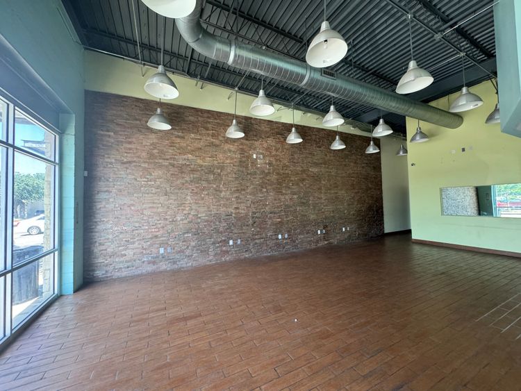 Natural light floods this large empty commercial space, highlighting the rustic brick wall and contemporary light fixtures.